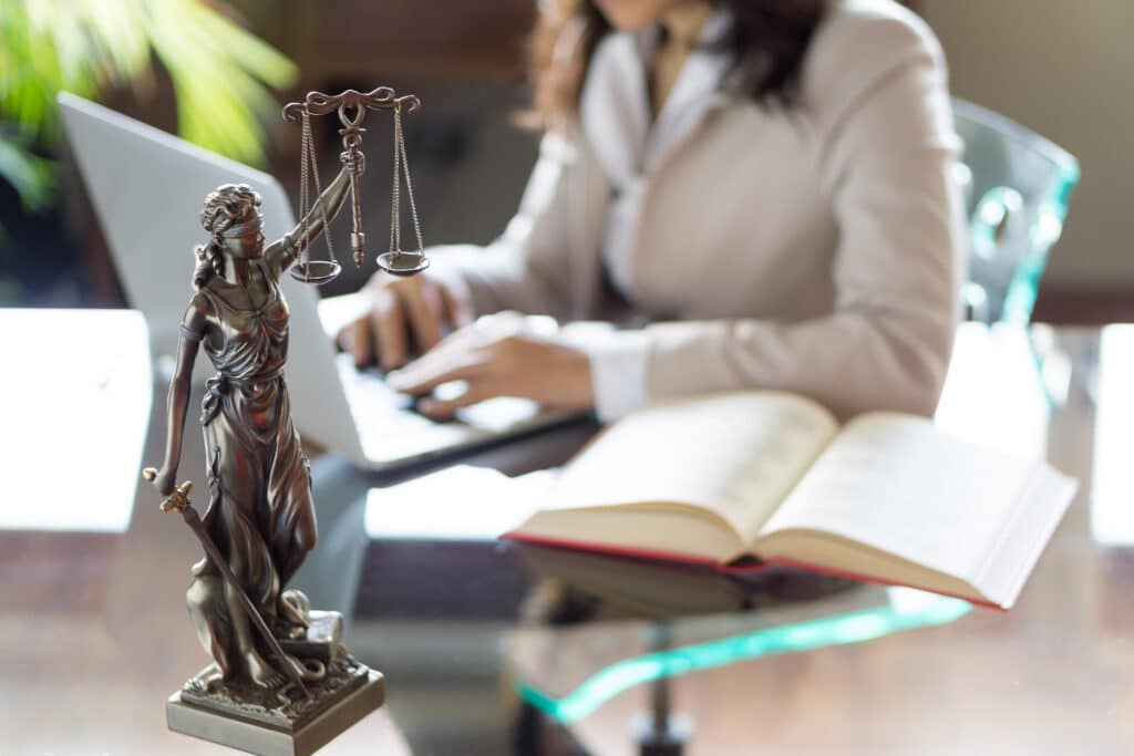 Statue de la Justice devant un avocat tapant sur un ordinateur portable, un livre ouvert sur un bureau.