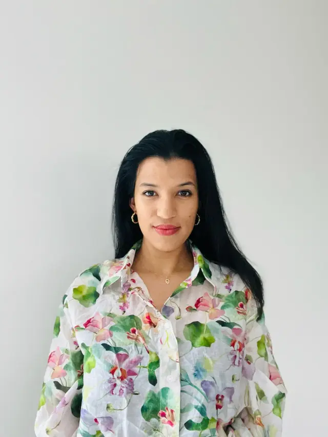 Portrait de femme aux cheveux noirs, souriant légèrement, portant une chemise florale blanche et transparente avec un fond blanc uni.